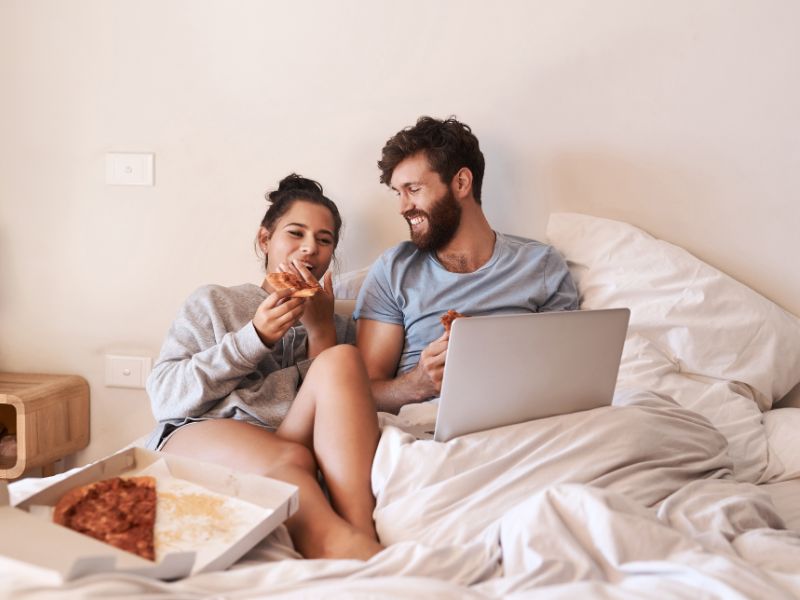 Couple in bed eating pizza and planning their wedding on a laptop